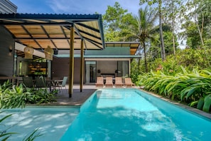 A modern outdoor pool area with a covered seating area and lush tropical vegetation.