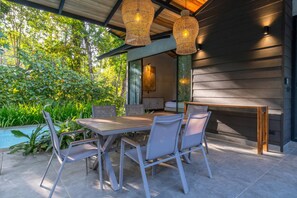 An outdoor patio with a dining table and chairs, surrounded by lush greenery and a pool.