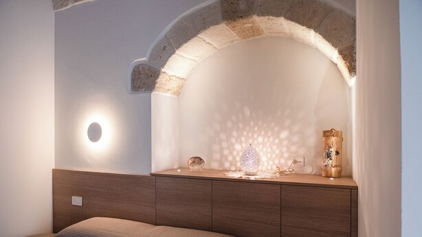 A bedroom with a bed, a nightstand, a decorative wall arch, and a wall-mounted light. - Pachita Seaside Rooms (Polignano a Mare)