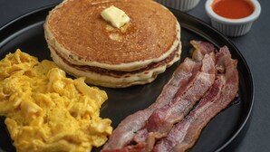 En frukosttallrik med pannkakor, äggröra, bacon och en skål med frukt.