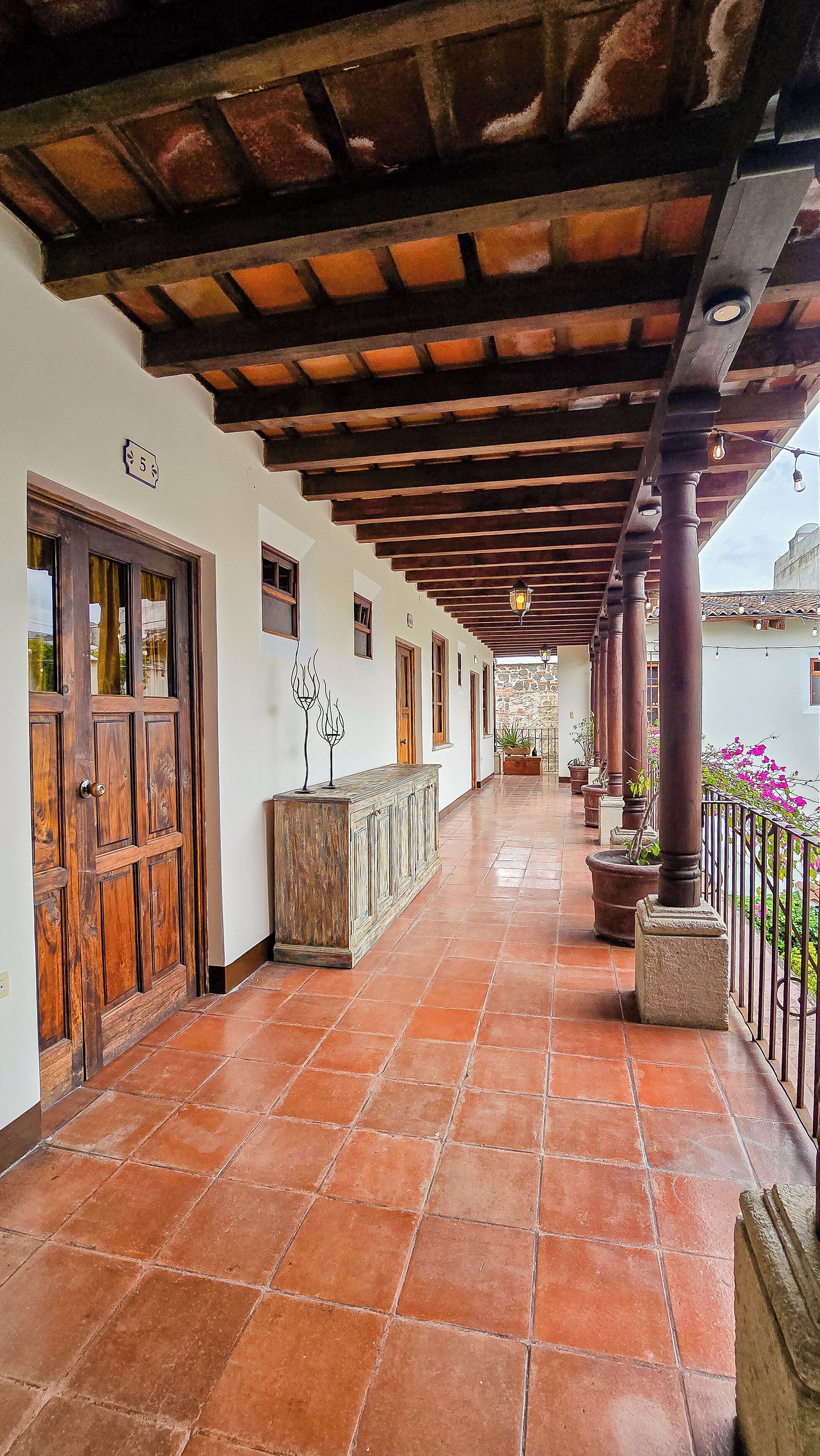 Un corredor cubierto con piso de baldosas, puertas de madera y columnas.