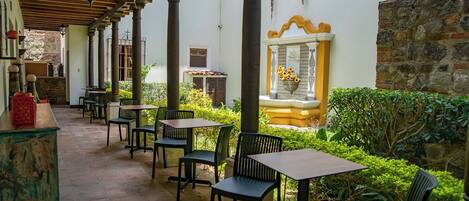 A covered outdoor seating area with tables and chairs, a stone wall, and a fountain with flowers.
