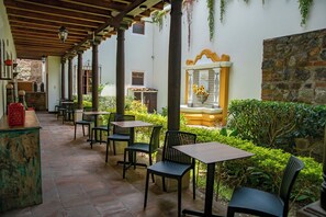 A covered outdoor seating area with tables and chairs, a stone wall, and a fountain with flowers. - Hotel Casa Colonial by AHS (Antigua Guatemala)