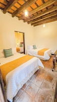 Habitación con tres camas, cada una con colcha blanca y almohada verde. El techo de madera presenta vigas a la vista y una lámpara colgante.
