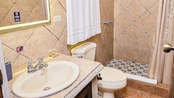 Un baño con lavamanos, inodoro y ducha. Hay un espejo sobre el lavamanos y una toalla colgada en un perchero.