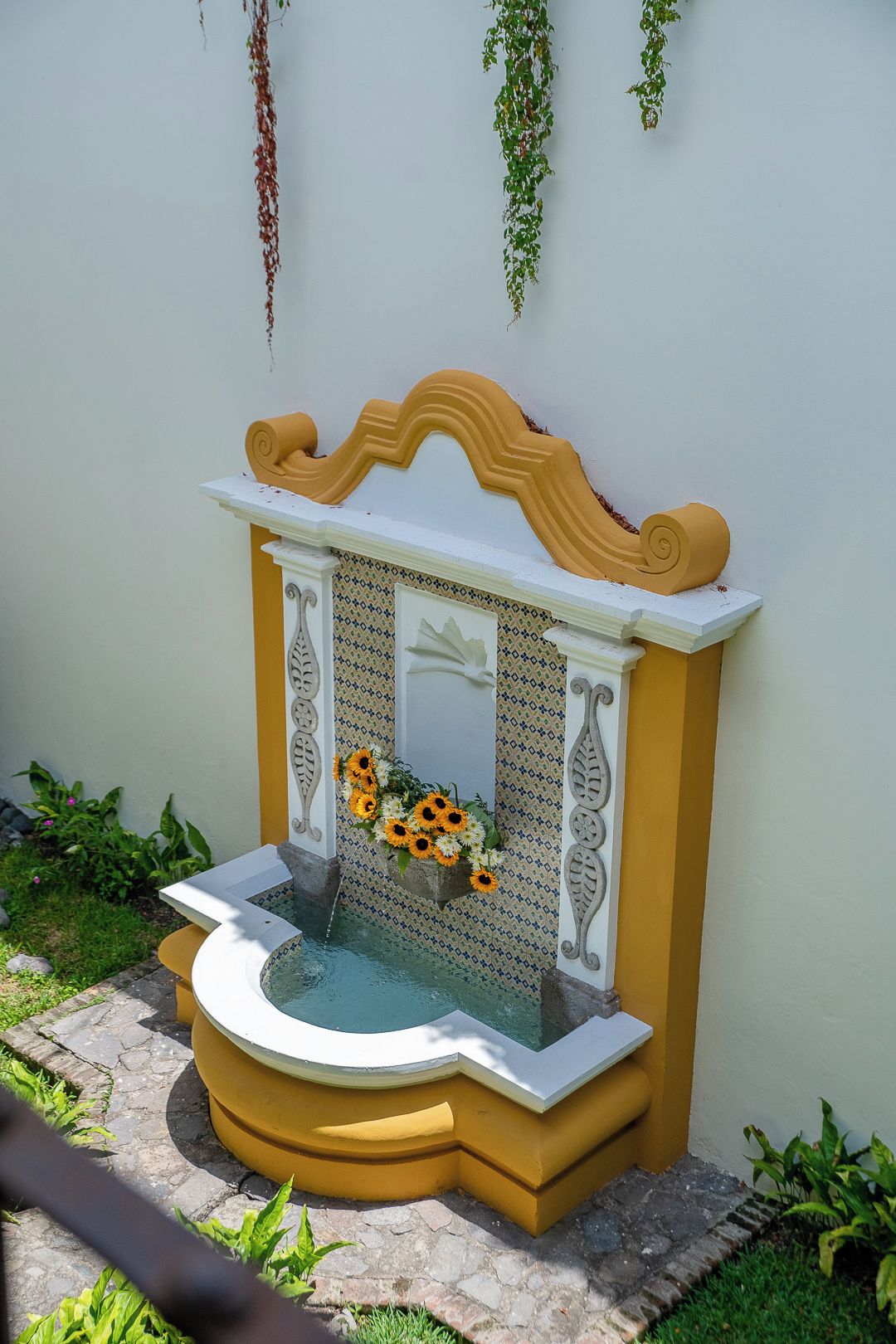 Una pequeña fuente con una parte superior decorativa, rodeada por un muro con diseño y una base amarilla.