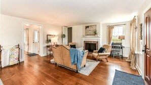 Ein geräumiges Wohnzimmer mit Holzboden, Kamin, einer Couch, einem Sessel und einem Essbereich.