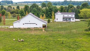 Ein weißes Scheunengebäude und ein weißes Haus mit Veranda, umgeben von einem grünen Feld und Bäumen.