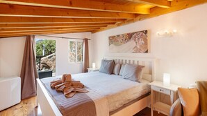 Ein Schlafzimmer mit Holzbalkendecke, einem Bett mit weißem Kopfteil, einem Nachttisch, einem Schreibtisch und einem Bild an der Wand.