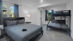 A bedroom with a large bed, a smaller bunk bed, a bedside table with a lamp, and a window with curtains.