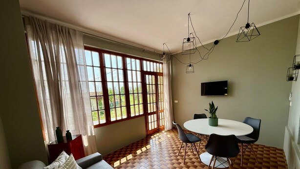 A living room with a sofa, a dining table with chairs, a TV mounted on the wall, and large windows with curtains. - ThreeZeroThree House • With Private Garden (Colombaro)