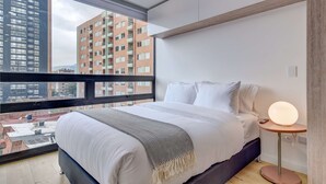 Moderni hotellihuone, jossa on suuri sänky, yöpöytä lampun kanssa ja suuri ikkuna, josta avautuu näkymä kaupungin rakennuksiin.