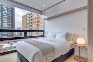 Habitació d'hotel moderna amb un llit gran, una taula de nit amb una làmpada, i vista a edificis de la ciutat a través d'un finestral ample.