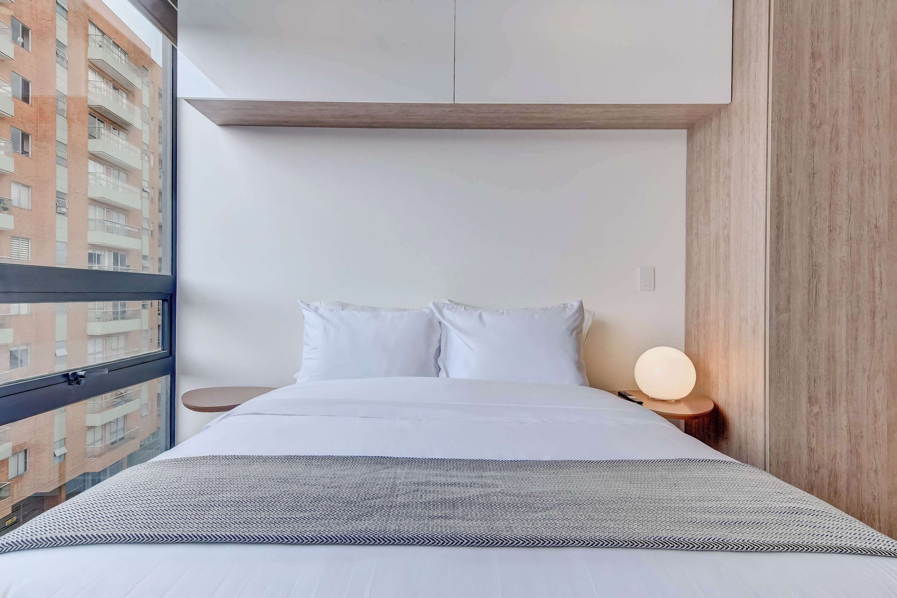 Une chambre d’hôtel moderne avec une grande fenêtre, un lit avec du linge de maison blanc, une table de chevet avec une lampe et une vue sur un immeuble.