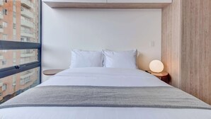 Moderni hotellihuone, jossa on suuri ikkuna, valkoisilla lakanoiden peitetty sänky, yöpöytä lampun kanssa ja näkymä rakennukseen.