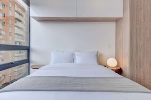 Habitació d'hotel moderna amb una finestra gran, un llit amb roba de llit blanca, una taula de nit amb una làmpada i vista a un edifici.