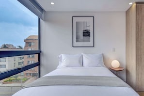 A modern bedroom with a large window, a framed picture on the wall, a bedside table with a lamp, and a bed with white bedding.