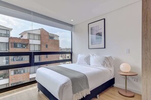 Une chambre avec une grande fenêtre, un lit, une table de chevet avec une lampe et un tableau encadré au mur.