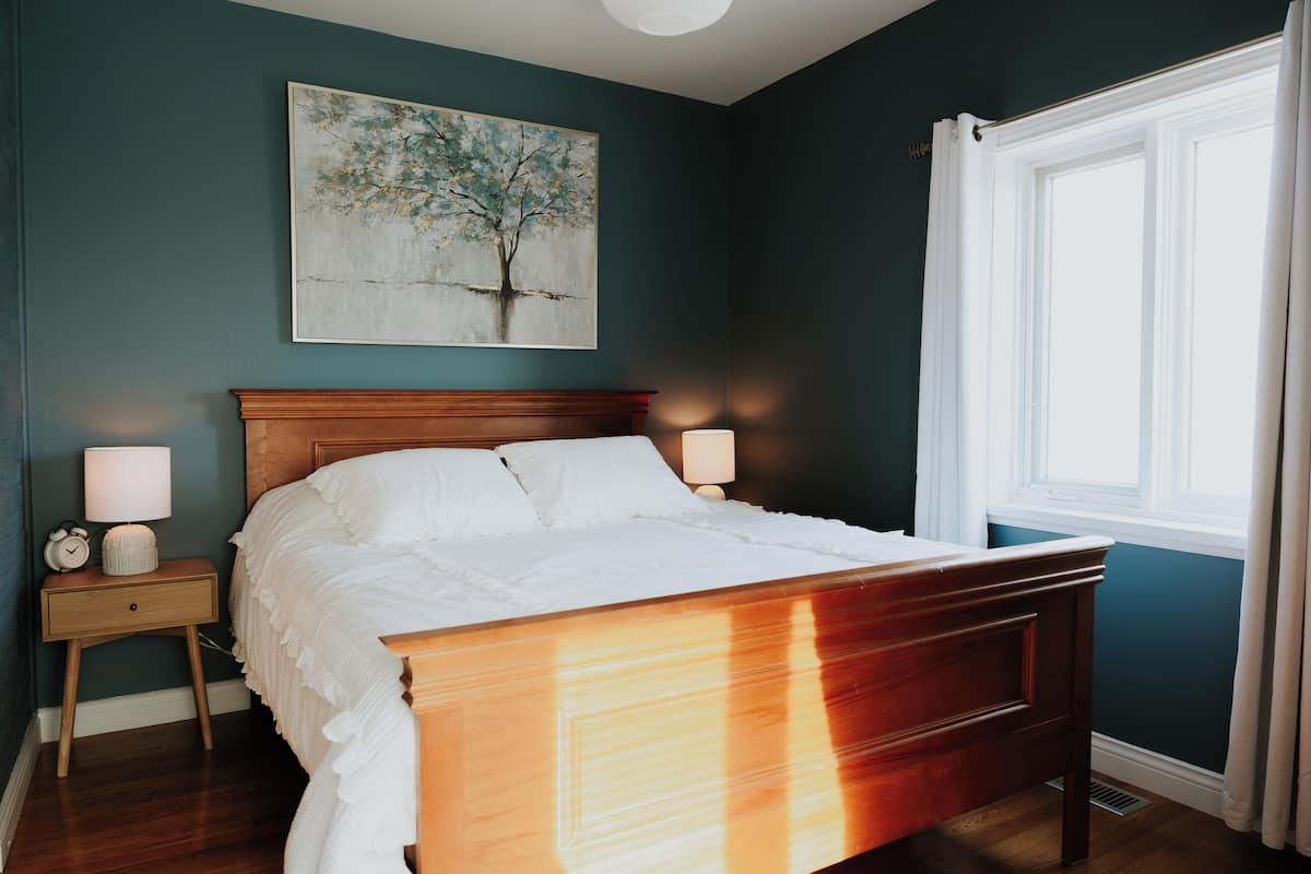 A bedroom with a wooden bed, a nightstand with a lamp, a wall-mounted painting, and a window with curtains.