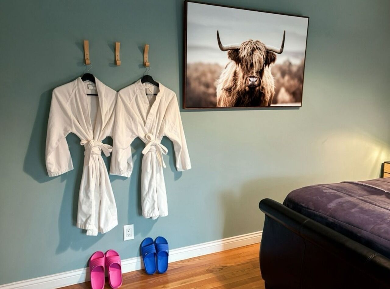 A bedroom with two robes hanging on a wall, a pair of slippers on the floor, and a bed with a purple cover.