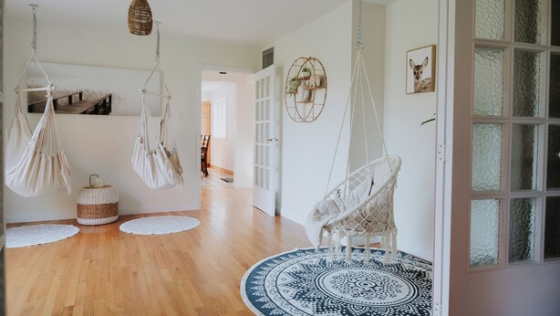 Una habitación con piso de madera, un tapete redondo con diseño, sillas colgantes de macramé, una cesta tejida y una lámpara colgante.