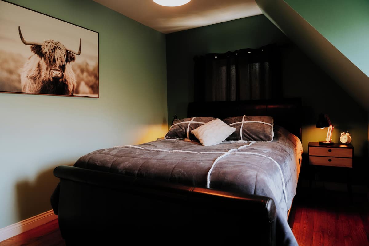 A bedroom with a bed, pillows, a nightstand with a lamp, a framed picture on the wall, and a sloped ceiling.