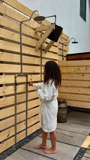 A child in a white bathrobe standing next to a wooden wall with metal pipes and a showerhead. - LE HAVRE BORÉAL (Grondines)