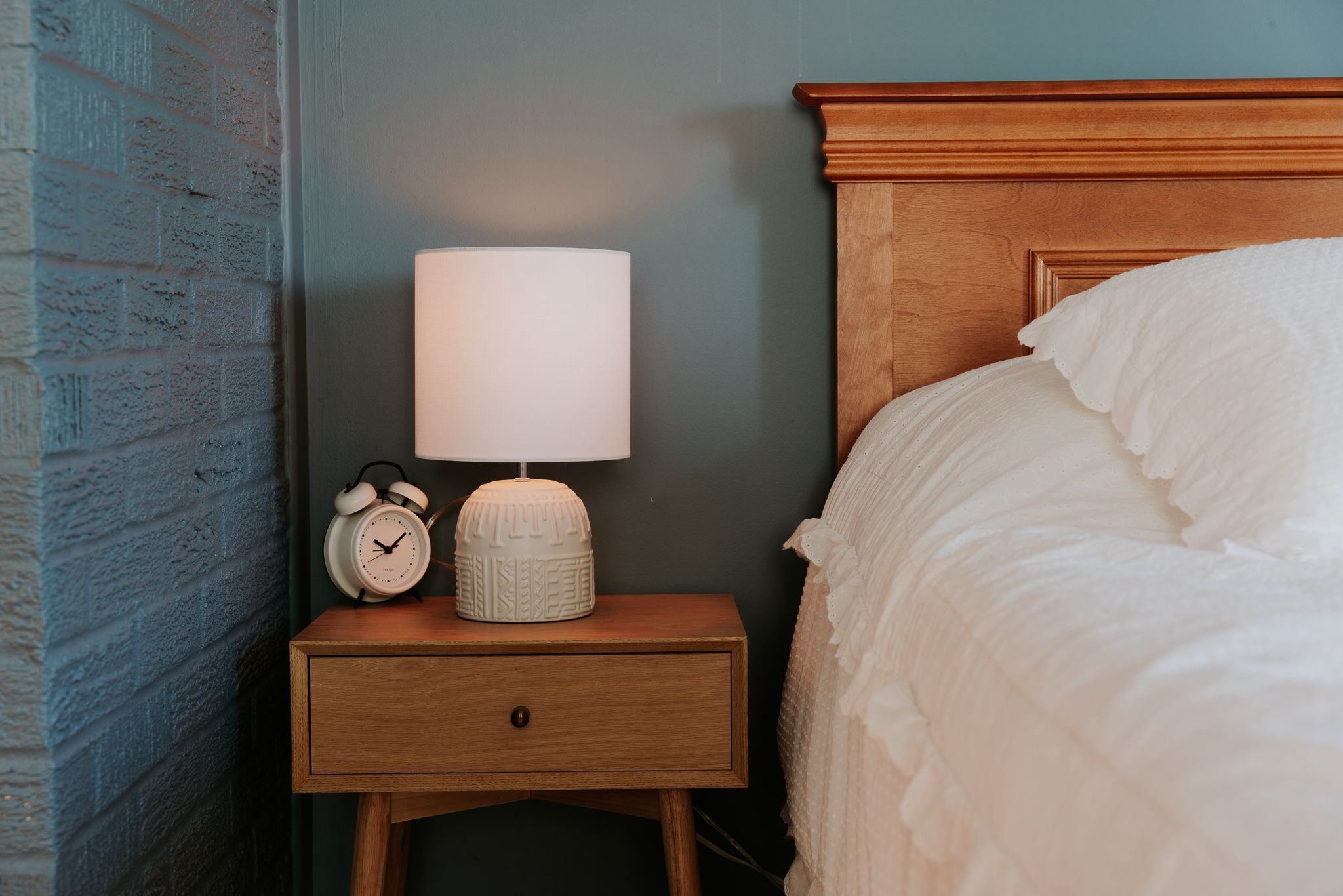 Una mesita de noche de madera con una lámpara, un reloj despertador y una cama con ropa blanca.