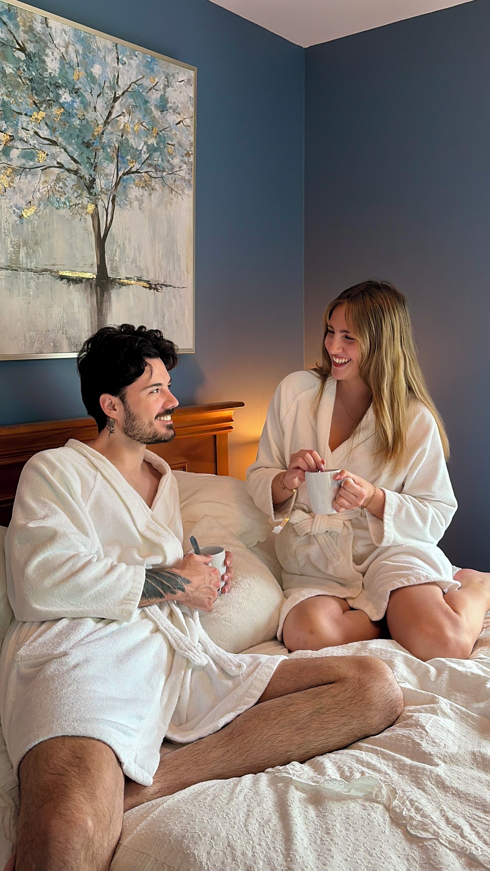 A couple in white bathrobes sitting on a bed with a painting of a tree on the wall.
