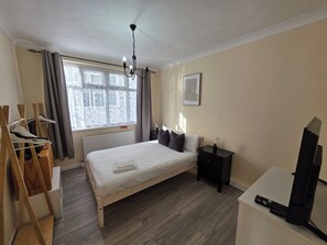 A bedroom with a bed, bedside table, a black TV stand, a window with curtains, and a wooden chair with hangers. - Modern Ground Floor Flat w Parking Romford (Greater London)