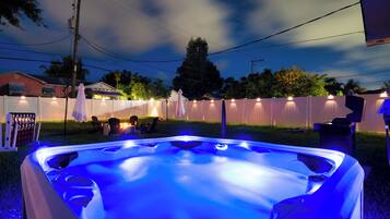 A hot tub with a blue light, surrounded by a white fence, chairs, and a fire pit.