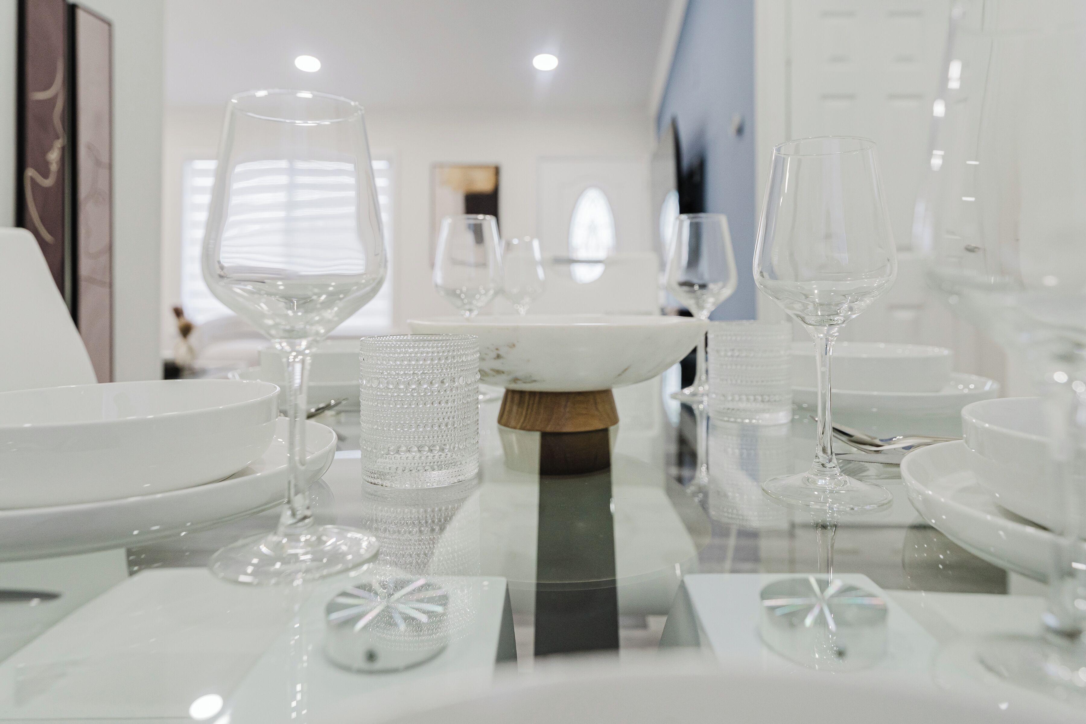 A dining table with clear glassware and white plates, set in a modern interior with a reflective surface.
