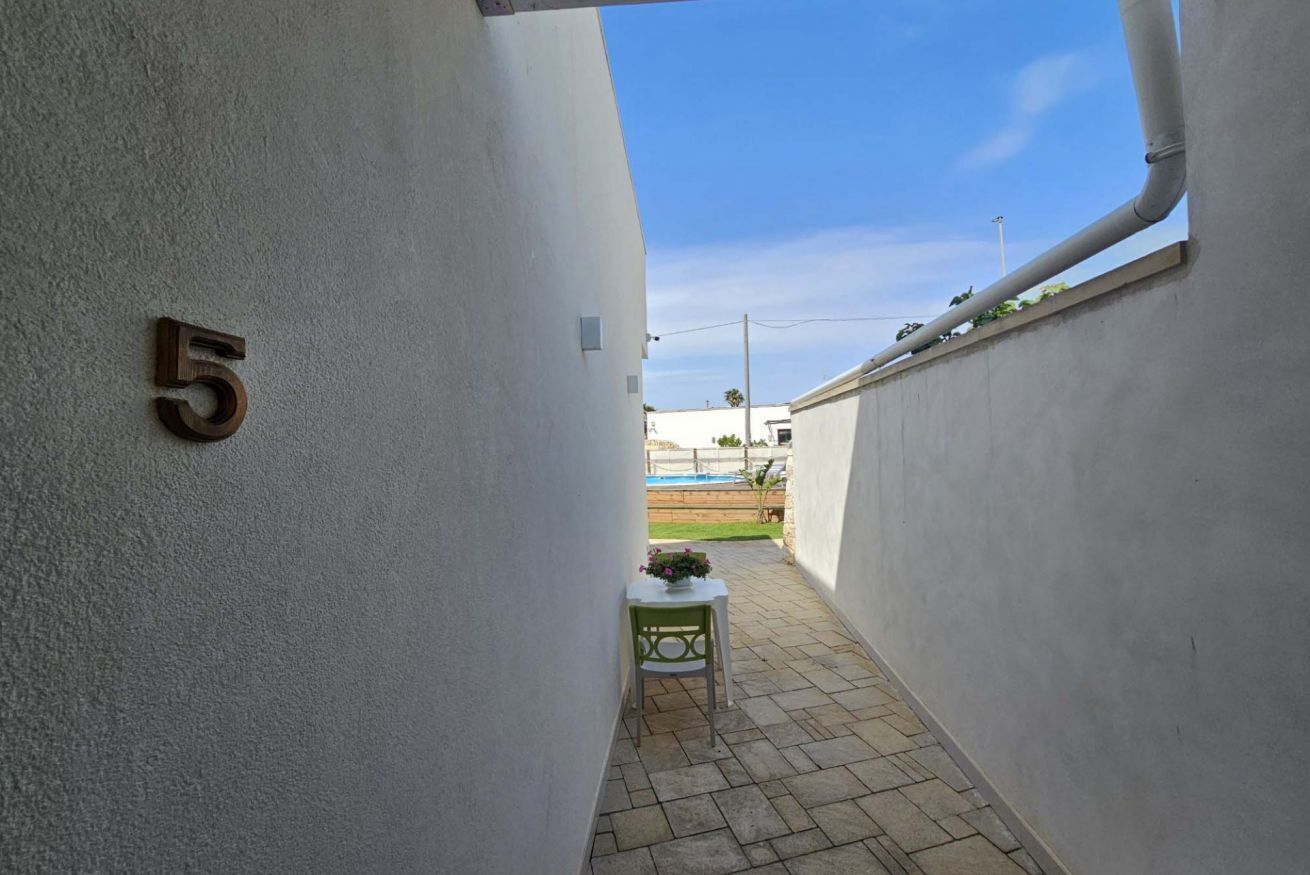 A narrow corridor with a number '5' on the wall, a small table with a flower vase, and a view of a pool and greenery in the distance.