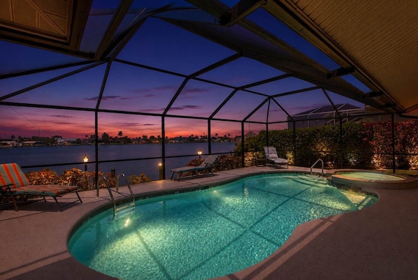 An indoor swimming pool with a glass ceiling, two lounge chairs, and a view of the sunset over water. - Cape Escape Home (jacuzzi) (Cape Coral)