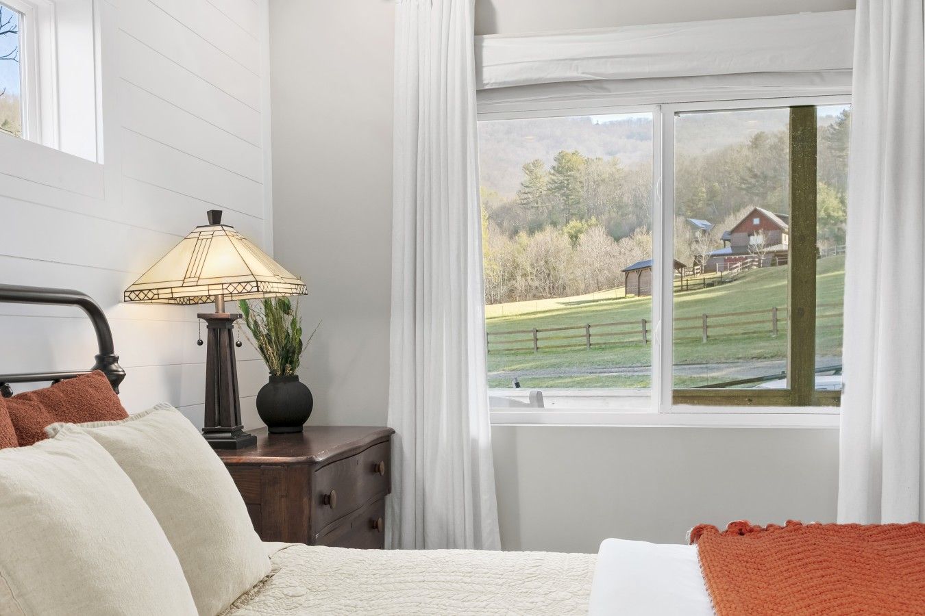 Un dormitorio con cama, una mesita de noche con lámpara y ventana con vista a un paisaje rural.