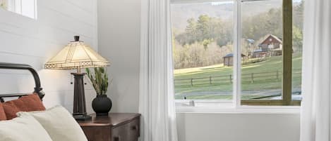 Un dormitorio con cama, una mesita de noche con lámpara y ventana con vista a un paisaje rural.