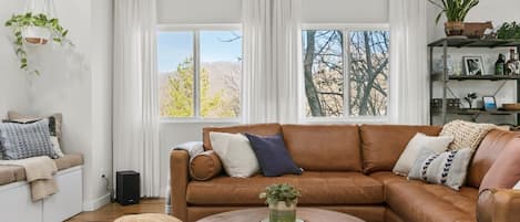 Ein Wohnzimmer mit braunem Ledersofa, einem runden Holztisch und einem weißen, an der Wand befestigten Regal mit Pflanzen.