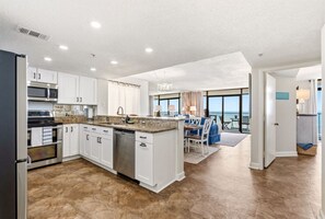 A modern kitchen with white cabinetry, stainless steel appliances, and a large island. There is a dining area with a table and chairs, and a view of the outdoors through large windows. - Oceanfront 4BR Condo | Stunning Views + Lazy River (North Myrtle Beach)