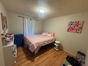 A bedroom with a bed, a bedside table, a small white stool, a wall-mounted painting, and a window with curtains. - "The Hangout" - Perfect for enjoying Tannenbaum Resort & Greers Ferry Lake! (Drasco)