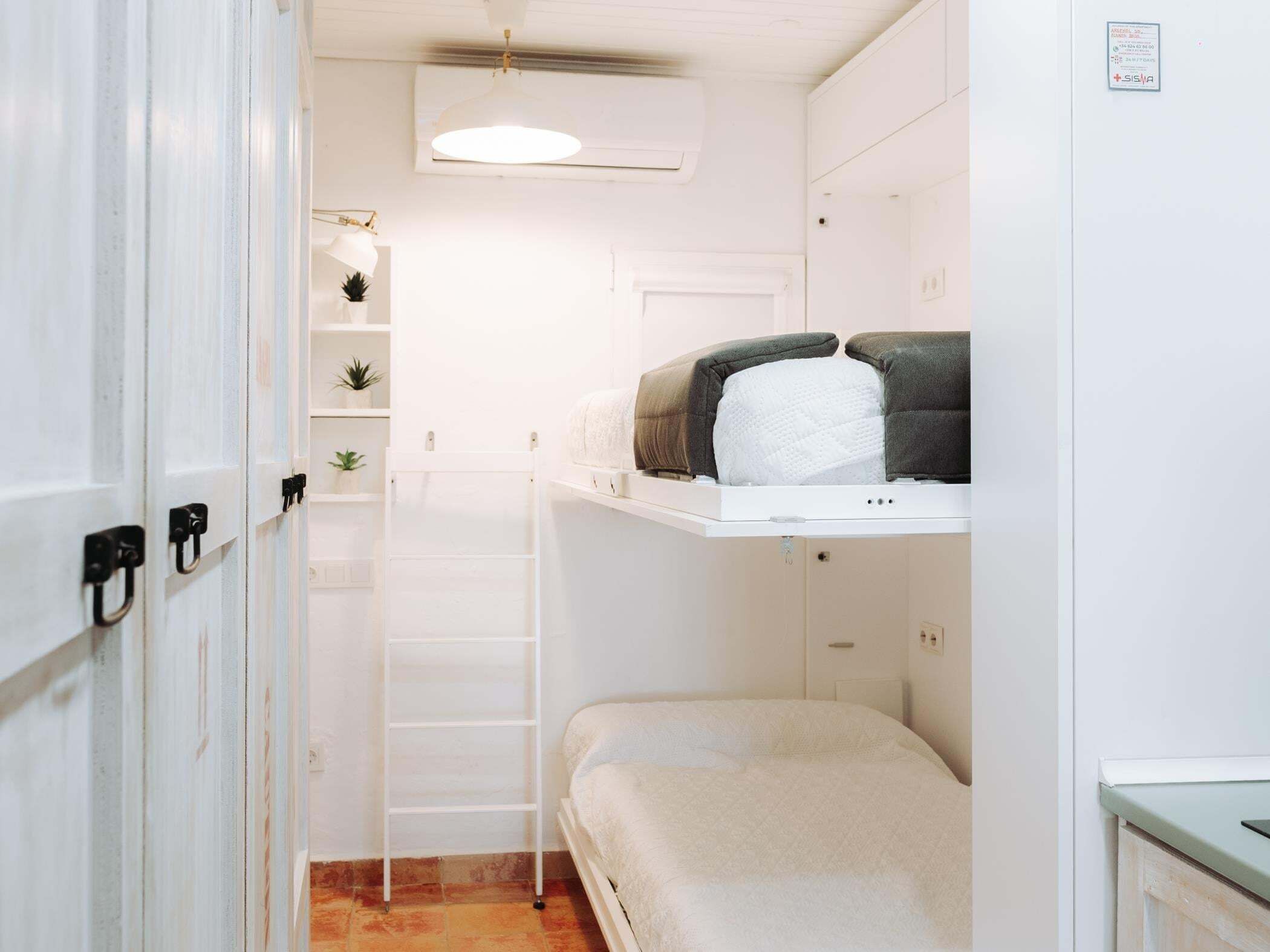 Een kleine kamer met een stapelbed, een klein plankje met planten, een witte deur en een plafondlamp.