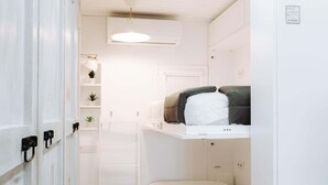 Ein kleines Zimmer mit Etagenbett, einer kleinen Ablagefläche mit Pflanzen, einer weißen Tür und einer Deckenleuchte.