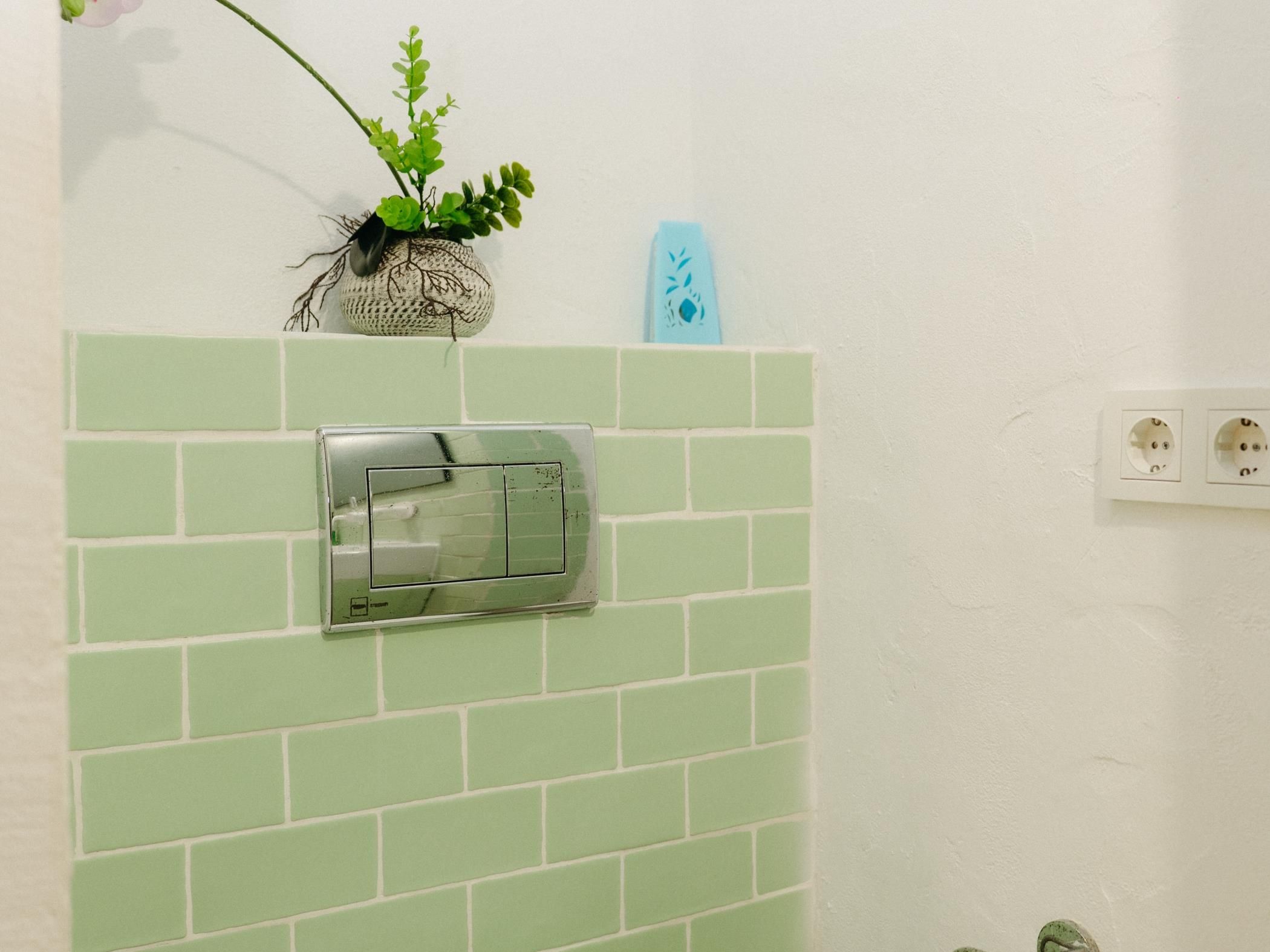 A bathroom with a green-tiled wall, a towel rack, a plant, and a blue soap dispenser.