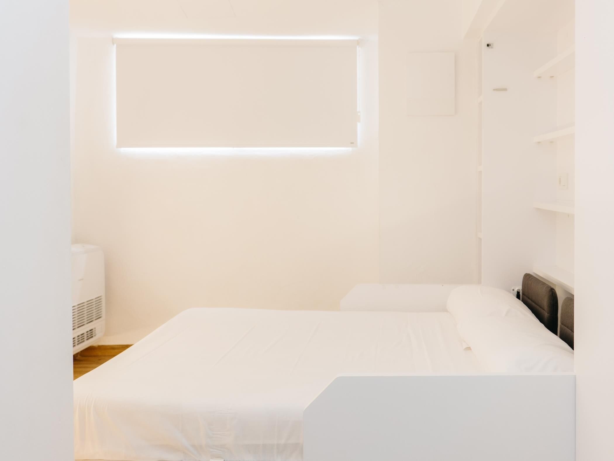 A minimalist bedroom with a bed, a window, and a wall-mounted shelf.