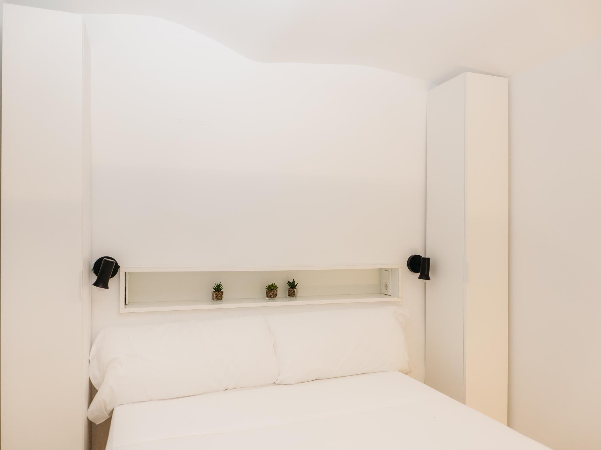 A minimalist bedroom with a single bed, a shelf with small plants, and a wall-mounted light fixture.