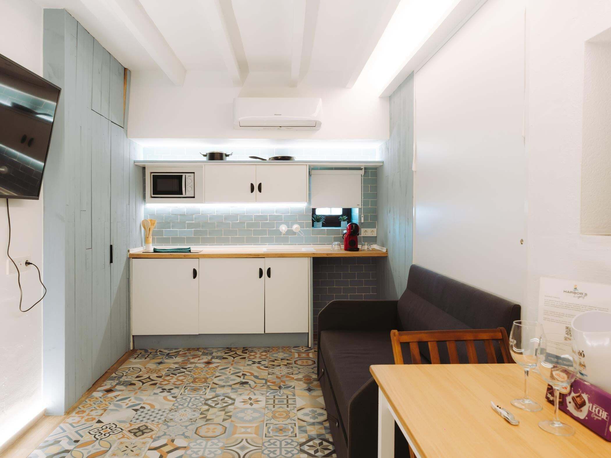 Una cocina compacta con microondas, un lavamanos pequeño y estufa. Hay un sofá de color oscuro y una mesa de madera con una caja de bombones y vasos.