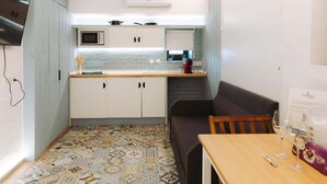 Una cocina compacta con microondas, un lavamanos pequeño y estufa. Hay un sofá de color oscuro y una mesa de madera con una caja de bombones y vasos.