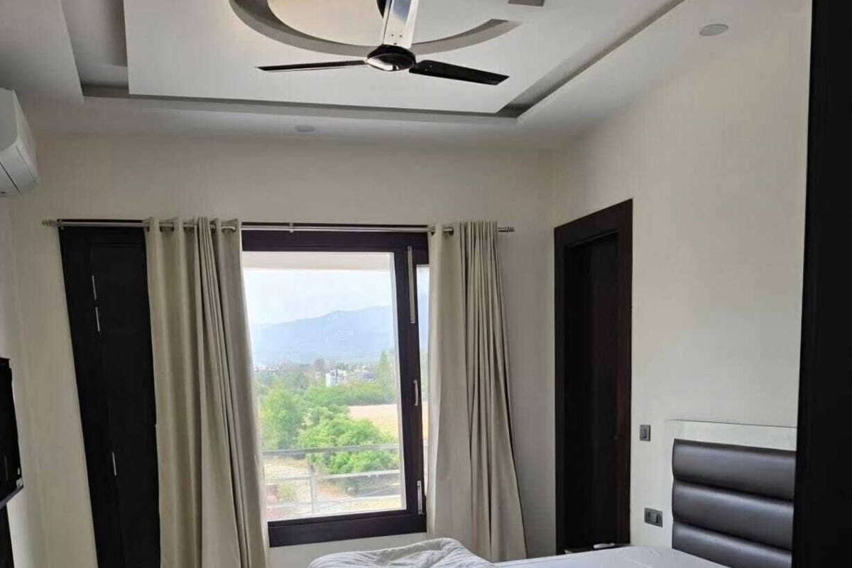 Una habitación de hotel moderna con una cama, un escritorio, una silla, un ventilador de techo y un ventanal con cortinas.