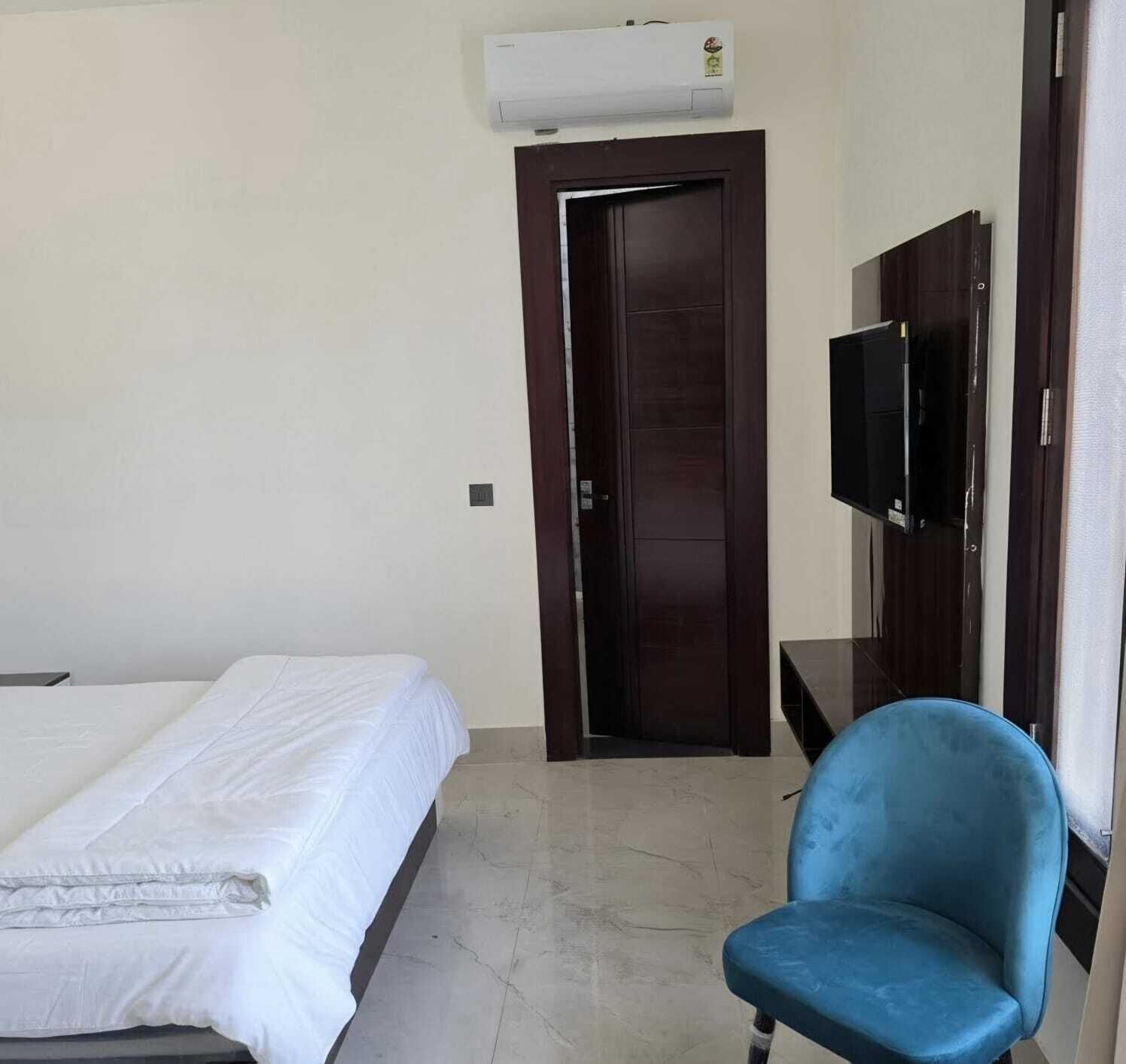 A hotel room with a bed, a blue chair, a TV mounted on the wall, and an air conditioning unit above the door.