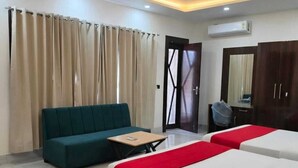 A hotel room with a sofa, two beds, a small table, a ceiling fan, and an air conditioning unit. - Hotel Golden Peak (Tanakpur)