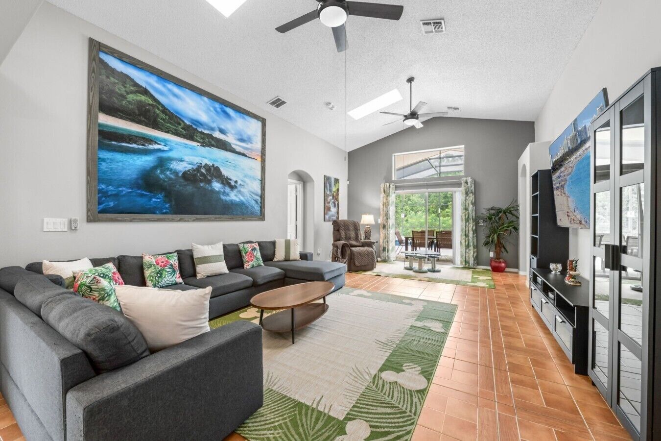 Een ruime woonkamer met een grote flatscreen-tv, een grijze hoekbank en een groot schilderij van een strandgezicht.